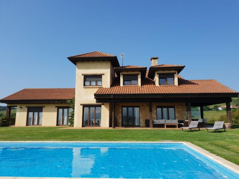 Casa de dos pisos con techo de tejas rojas y grandes ventanales, situada en un césped con una piscina rectangular en primer plano bajo un cielo azul claro.