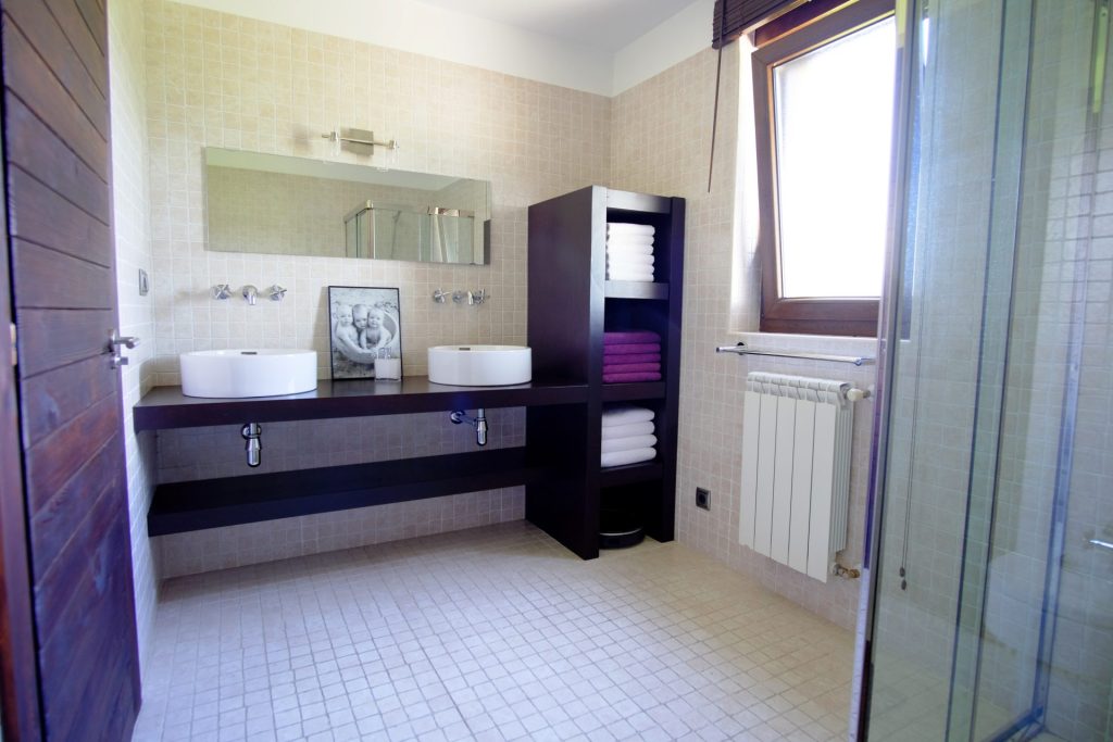 Baño moderno con azulejos beige, dos lavabos blancos sobre un mueble de madera oscura y estantes con toallas perfectamente apiladas. Un gran espejo y una ventana aportan luminosidad.