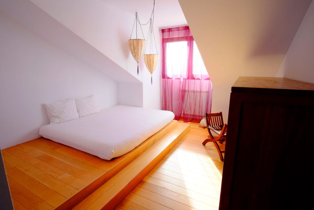 Un dormitorio minimalista con una cama baja tipo plataforma, ropa de cama blanca y dos almohadas. Una luz tenue se filtra a través de unas cortinas rosas translúcidas. Tres lámparas colgantes de mimbre crean un ambiente acogedor.