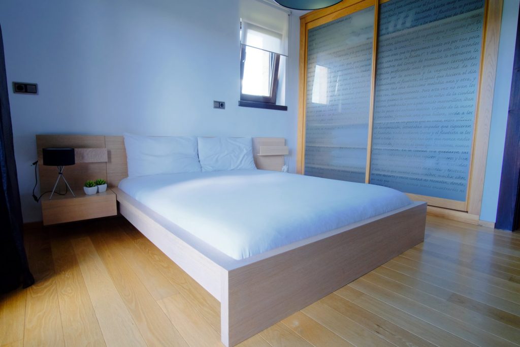 Dormitorio minimalista con suelo de madera, cama baja con ropa de cama blanca, mesitas de noche de madera y puerta corredera con texto. Ambiente luminoso y sereno.