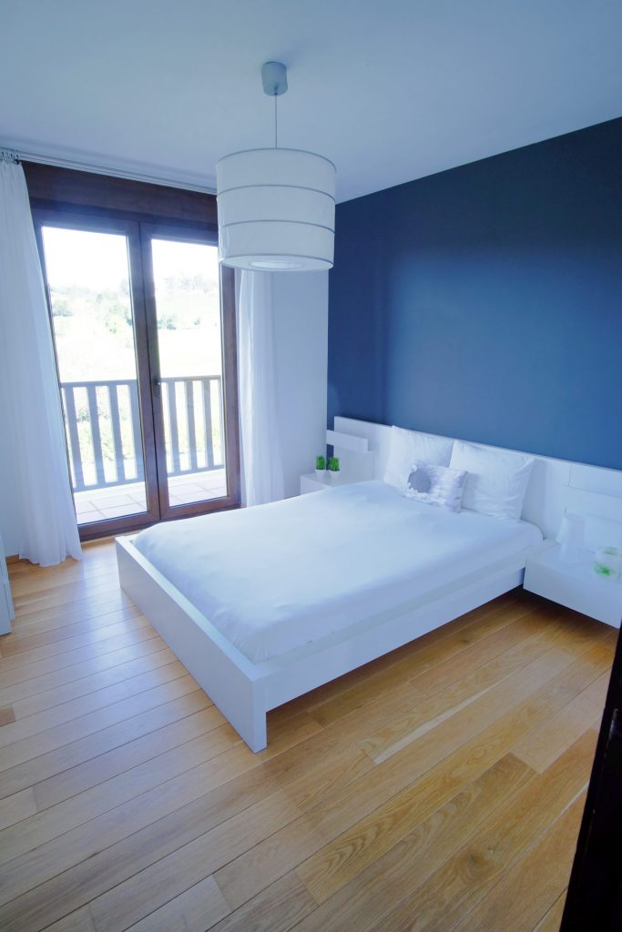 Un dormitorio minimalista con cama blanca, suelo de madera y una pared azul oscuro. Grandes ventanales con cortinas traslúcidas dejan entrar la luz natural, creando una atmósfera serena.