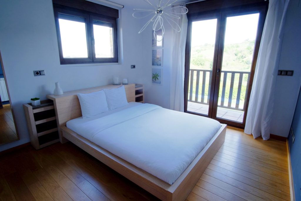 Dormitorio moderno con suelo de madera, cama minimalista con ropa de cama blanca, gran ventanal con cortinas vaporosas y vistas al balcón. Ambiente luminoso y sereno.