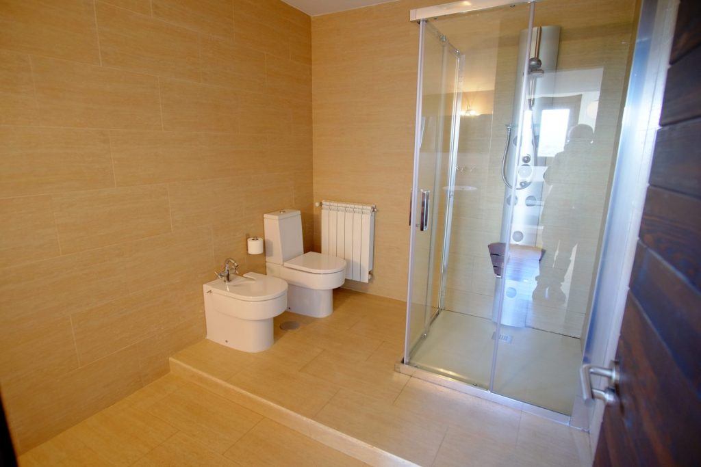 Un baño moderno con paredes y suelo de azulejos color beige. Cuenta con bidé, inodoro y una ducha con mampara de cristal de diseño elegante y grifería metálica.