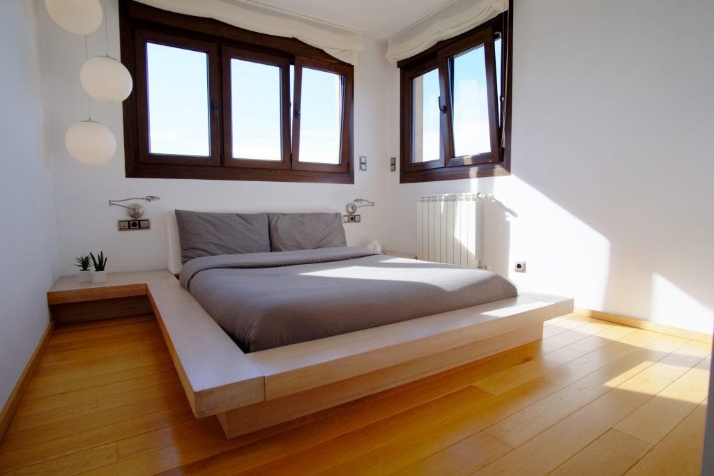 Dormitorio minimalista con cama baja tipo plataforma, ropa de cama gris y suelo de madera clara. Grandes ventanales con marcos de madera permiten la entrada de abundante luz natural. Ambiente acogedor y sereno.