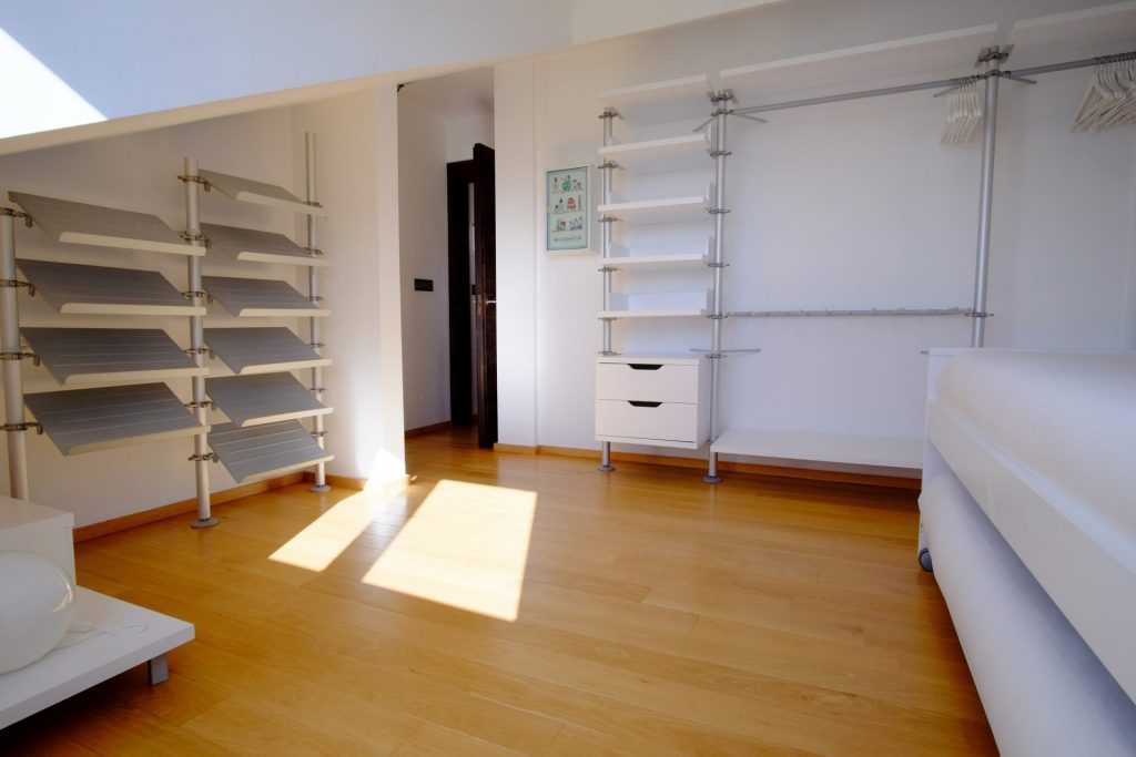 Habitación luminosa y minimalista con suelo de madera. Estanterías y perchas metálicas vacías adornan las paredes blancas. Un rayo de sol se filtra por el suelo. Ambiente tranquilo y ordenado.