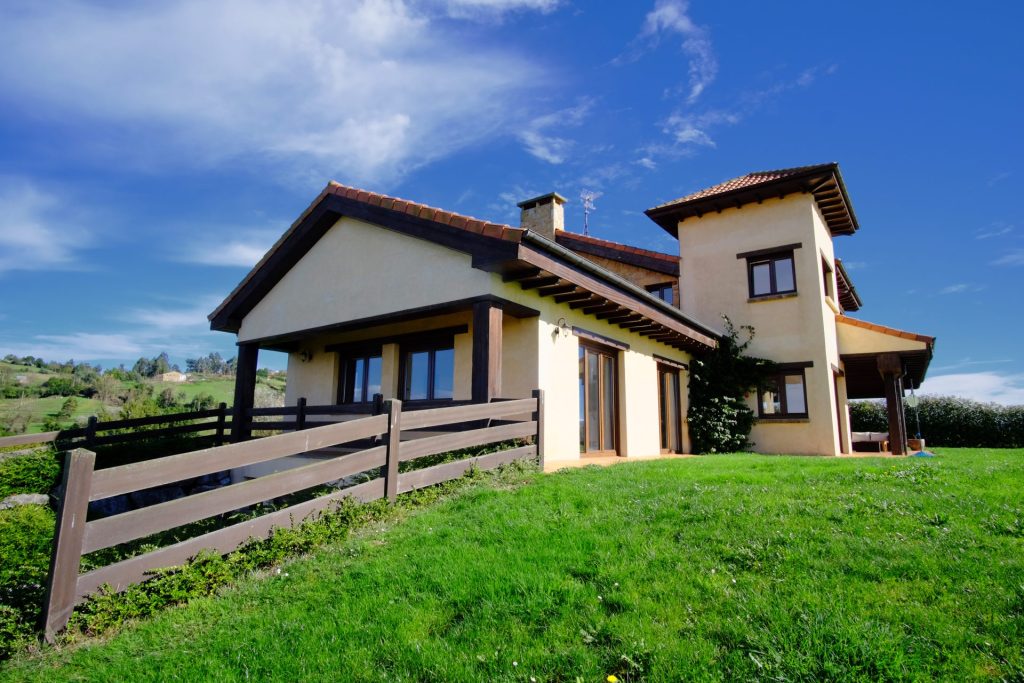 Una pintoresca casa de campo con un tejado inclinado de tejas rojas, enclavada en una exuberante colina verde. Cuenta con una valla de madera y se recorta contra un cielo azul despejado.
