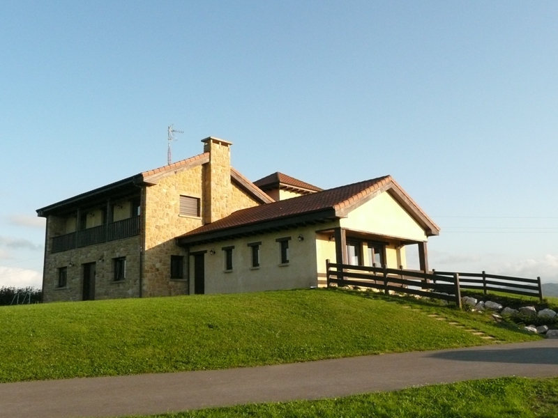 Una casa de dos plantas, de piedra y estuco, con tejado de tejas rojas, se alza sobre una colina cubierta de hierba junto a una carretera asfaltada bajo un cielo despejado.