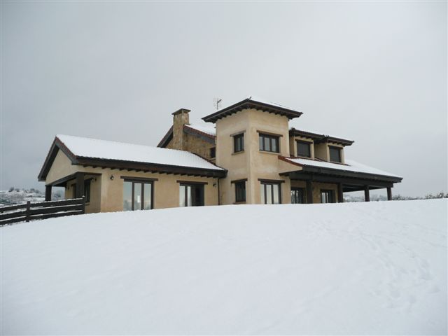 Una casa moderna, de color beige y con grandes ventanales, se encuentra rodeada de un paisaje nevado bajo un cielo gris y nublado, creando una atmósfera serena e invernal.
