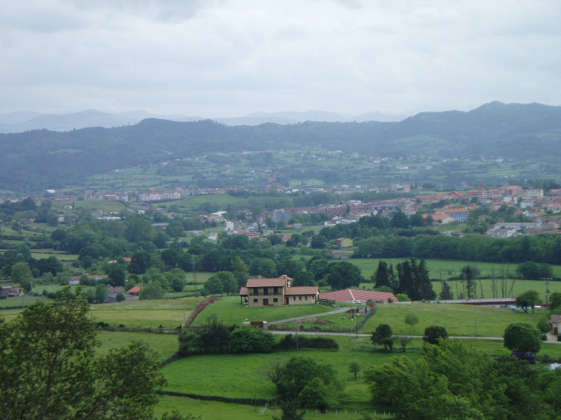 Un paisaje rural con una gran casa en primer plano, rodeada de exuberantes campos verdes y árboles, y un pueblo lejano bajo un cielo nublado.